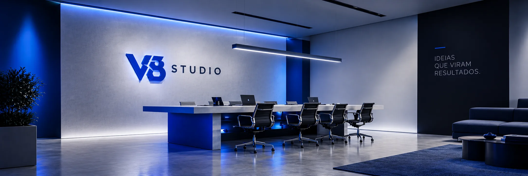 Espaço de trabalho com o logo V8 Studio iluminado na parede, mesa longa com notebooks e luz azul.