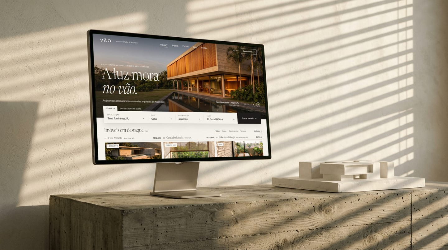 Computador sobre um bloco de concreto, com sombras de brise na parede, mostrando o site do estúdio fictício VÃO com uma casa na hora dourada.